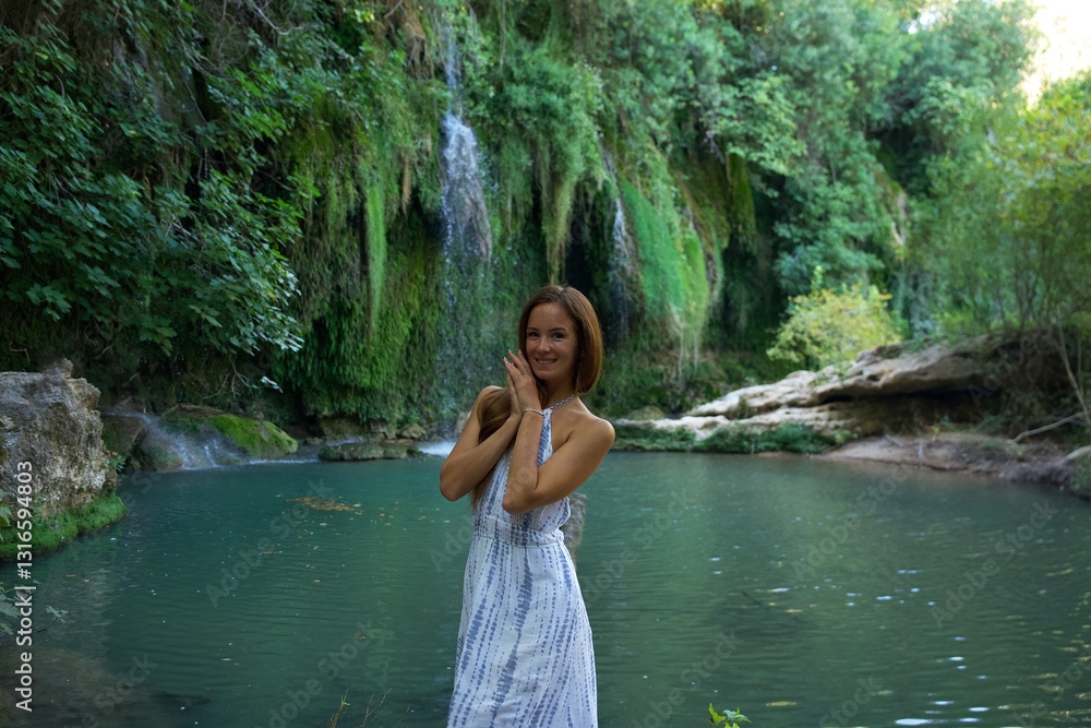 Fototapeta premium A joyful woman in a white dress stands near a turquoise river with a waterfall in the background, surrounded by lush greenery, expressing happiness and serenity. 