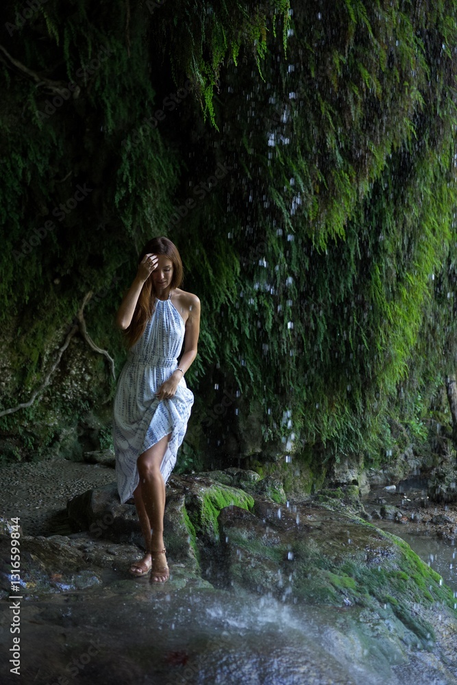 Fototapeta premium A woman in a white dress stands near a cascading waterfall, touching her hair as she enjoys the refreshing mist, surrounded by lush greenery and moss-covered rocks. 
