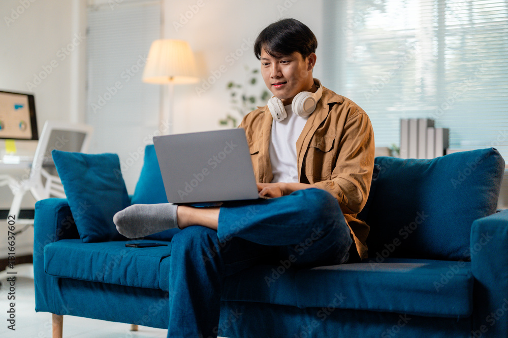 Young man using laptop and headphones while working remotely from home, sitting comfortably on sofa Young man using laptop and headphones while working remotely from home, sitting comfortably on sofa