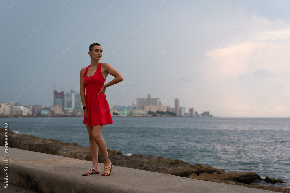 Young woman wearing vibrant red dress standing on malecon, posing against havana skyline with dramatic cloudy skies and ocean backdrop Young woman wearing vibrant red dress standing on malecon, posing against havana skyline with dramatic cloudy skies and ocean backdrop