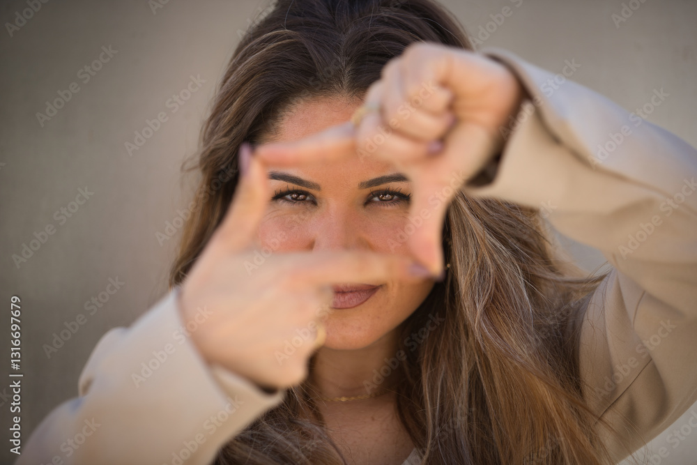 Close-up portrait of a young, beautiful, brown-haired woman in a light grey jacket suit, looking at the camera through a frame she forms with her hands, on a grey metallic background. Close-up portrait of a young, beautiful, brown-haired woman in a light grey jacket suit, looking at the camera through a frame she forms with her hands, on a grey metallic background.
