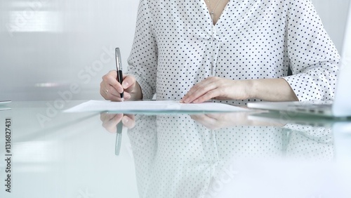 Businesswoman in white dotted blouse is signing a contract at a sleek glass desk in a modern office, showcasing professionalism and focus in her work environment. Business people concept