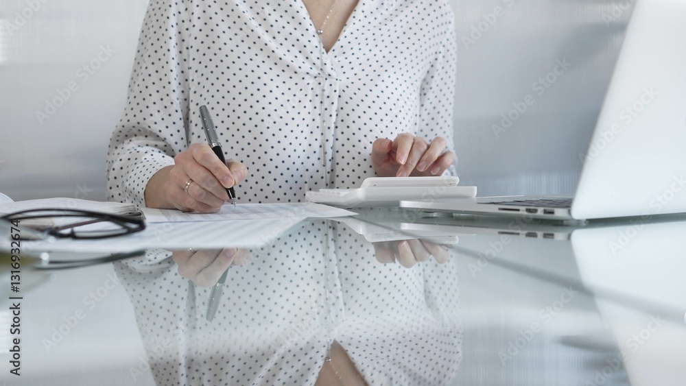 Unknown businesswoman using a calculator to crunch numbers and jotting down meticulous notes at a sleek glass desk in a contemporary office, closeup view. Taxes, audit in business