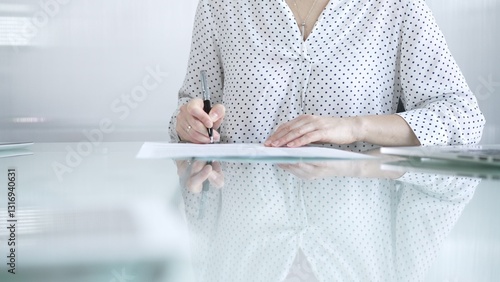 Businesswoman in white dotted blouse is signing a contract at a sleek glass desk in a modern office, showcasing professionalism and focus in her work environment. Business people concept