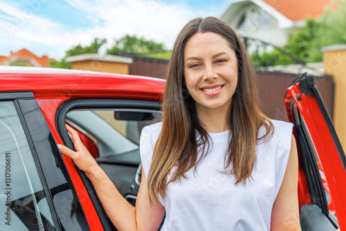 Obraz na płótnie A smiling brunette woman in a white shirt and black jeans stands next to a red car with the door open, enjoying the fresh air and scenic countryside on a sunny day. w salonie