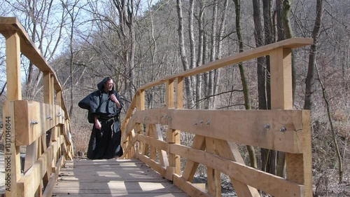 A man in black hooded monk's robes walks on a wooden bridge amidst a forest of bare trees. 