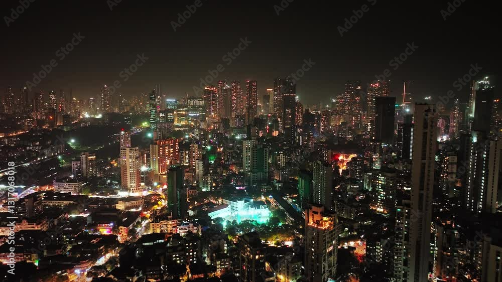 An aerial view of Mumbai's high-rise buildings and beautiful nightlights. The sky is cloudy, and there is medium traffic on the road. The cityscape is filled with modern illuminated buildings.