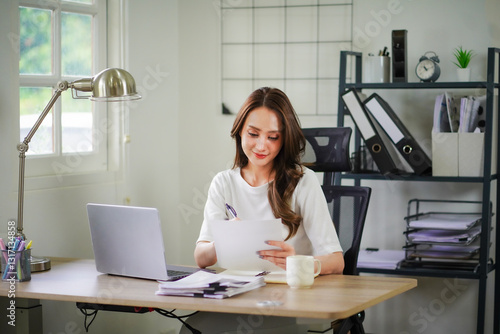 A woman is sitting at a desk with a laptop and a piece of paper in front of her