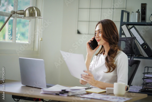 A woman is talking on her cell phone while sitting at a desk with a laptop