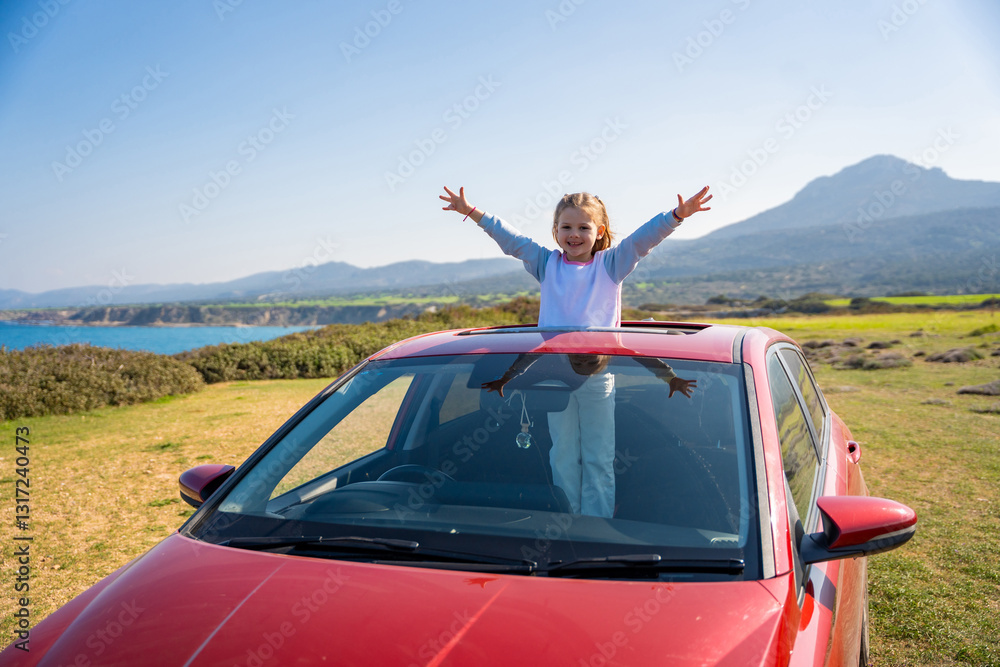 Fototapeta premium Little girl joyfully sticks out of car sunroof with raised hands against the backdrop of mountains in Northern Cyprus. The image symbolizes freedom, adventure, and the excitement of travel with child