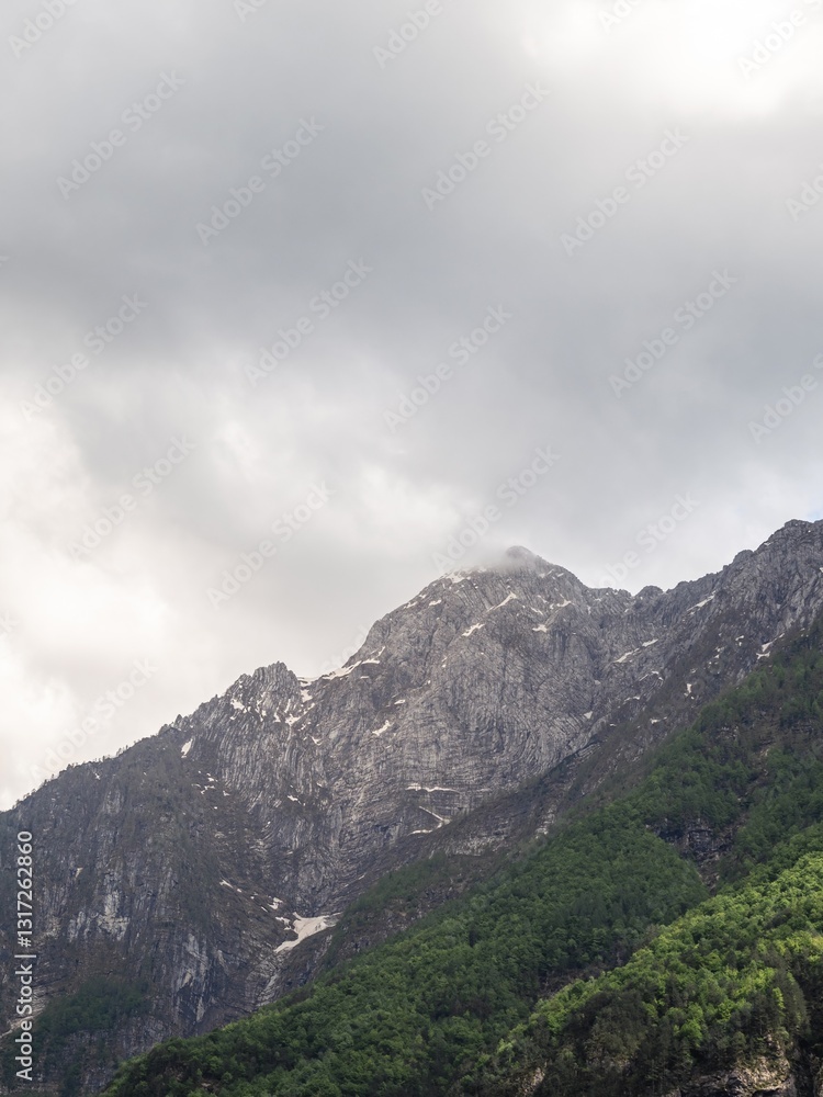 A towering mountain peak with rugged rock formations and patches of greenery, partially covered by clouds. The overcast sky adds a dramatic and moody atmosphere to the scene. A towering mountain peak with rugged rock formations and patches of greenery, partially covered by clouds. The overcast sky adds a dramatic and moody atmosphere to the scene.