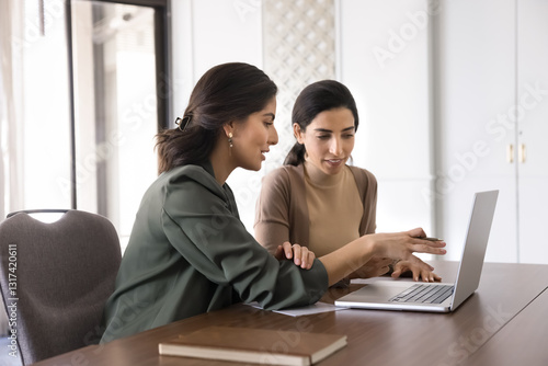 Canvas-taulu Two young women sit at desk reviewing report, document, or presentation on lapto