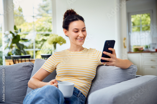 Happy woman using mobile phone  at home having coffe on sofa