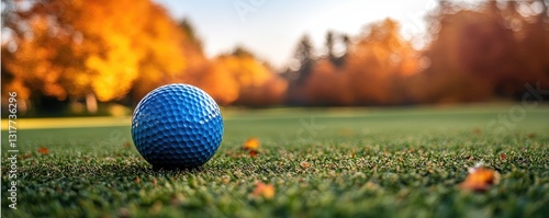 A blue golf ball rests on lush green grass in an autumn park setting, surrounded by vibrant orange and yellow leaves creating a warm, inviting atmosphere that features trees in fall colors