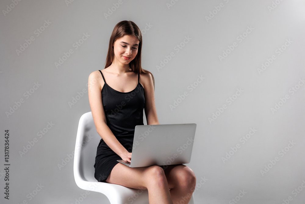 Fototapeta premium Young woman working on a laptop while sitting on a chair in a minimalist indoor space during the day
