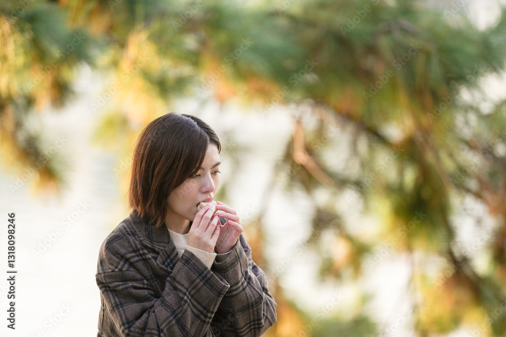 Winter at Ohori Park in Chuo Ward, Fukuoka City, Japan. A 29-year-old Japanese woman wearing a checkered coat. She sits on a chair by the pond, eating sweets. Winter at Ohori Park in Chuo Ward, Fukuoka City, Japan. A 29-year-old Japanese woman wearing a checkered coat. She sits on a chair by the pond, eating sweets.
