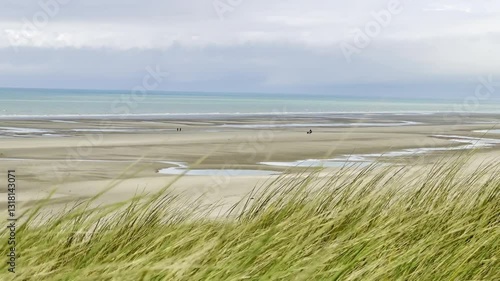 Serene Authie Bay coastal landscape with golden dune grass in the foreground, vast sandy beach, tidal pools, and a lone figure walking by the calm sea under a partly cloudy sky, near Fort-Mahon