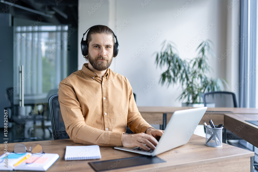 Obraz premium A handsome bearded man with headphones works on his laptop in a modern office setting, enhancing his work with music and focus.