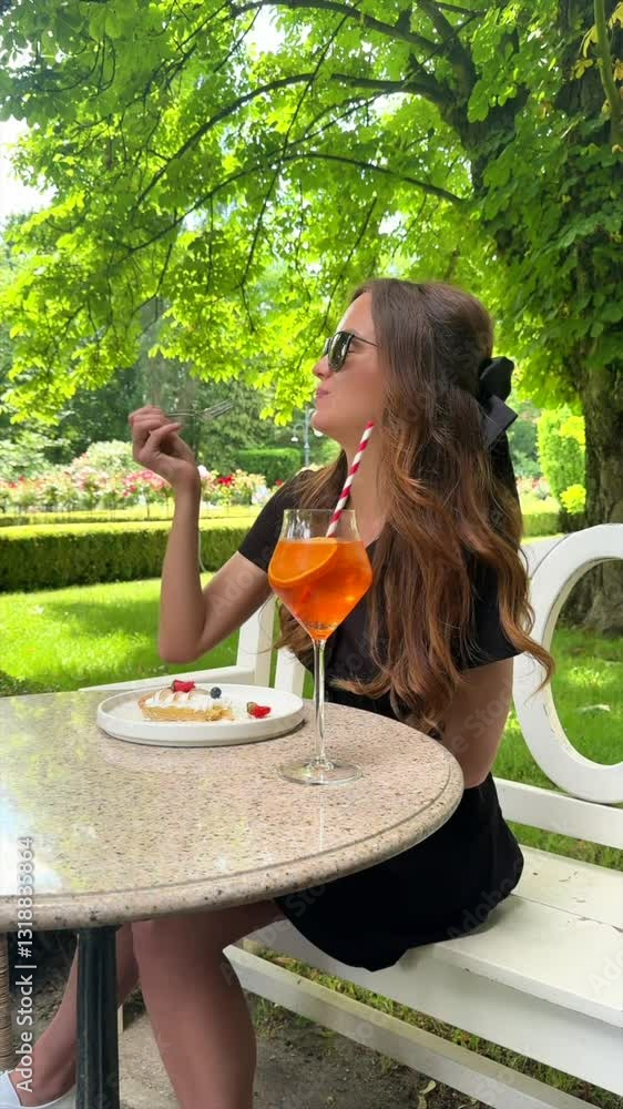 A beautiful woman in a black dress sits at a table in a park, savoring an Aperol cocktail and indulging in a delicious cake.