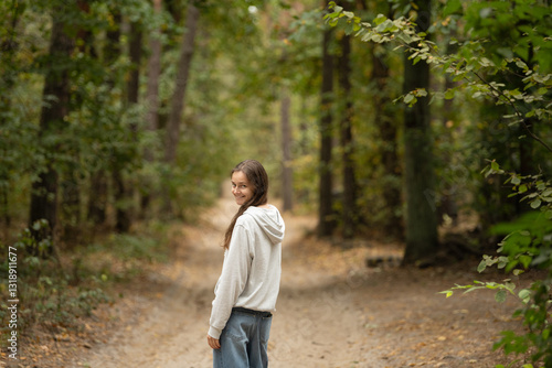 A young woman strolls along a dirt path in a lush green forest, wearing casual clothes. She glances back with a friendly smile, surrounded by tall trees and soft foliage