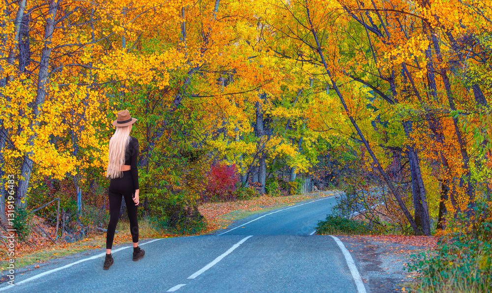 A young woman walks on an asphalt road, autumn trees on either side of the road