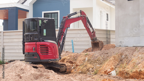 A small red excavator is digging a hole to prepare for building a wall around a concrete house on background Concept of working with machinery at a construction site.