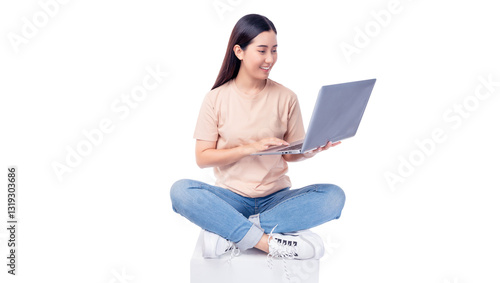 Cheerful young asian woman sitting cross-legged on a white cube while using a laptop. She wears T-shirt and blue jeans, smiling as she interacts with the mobile phone isolated on white background