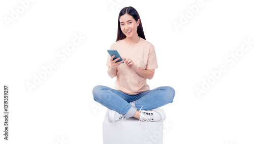 Cheerful young Asian woman sitting cross-legged on a white cube, engaging with smartphone. She wears T-shirt and blue jeans, smiling while interacting with the phone isolated on white background