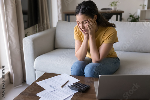 Brazilian woman sits at table with papers and laptop, feels stressed due to unpaid expenses, budgeting concerns, looks overwhelmed by debt, loans, or huge household utility bills, unexpected charges