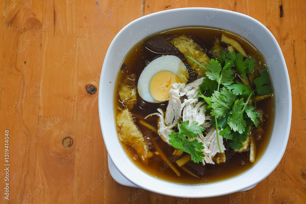 bowl of soup sits on a wooden table. The soup contains ingredients such as halved boiled eggs, shredded chicken, fresh coriander leaves, and what looks like bamboo shoots or other vegetables. bowl of soup sits on a wooden table. The soup contains ingredients such as halved boiled eggs, shredded chicken, fresh coriander leaves, and what looks like bamboo shoots or other vegetables.