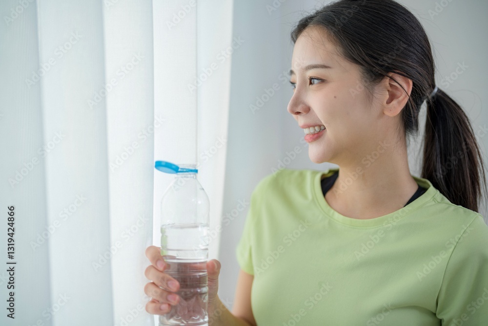 Woman in green shirt smiles while holding water bottle near window during bright morning Woman in green shirt smiles while holding water bottle near window during bright morning