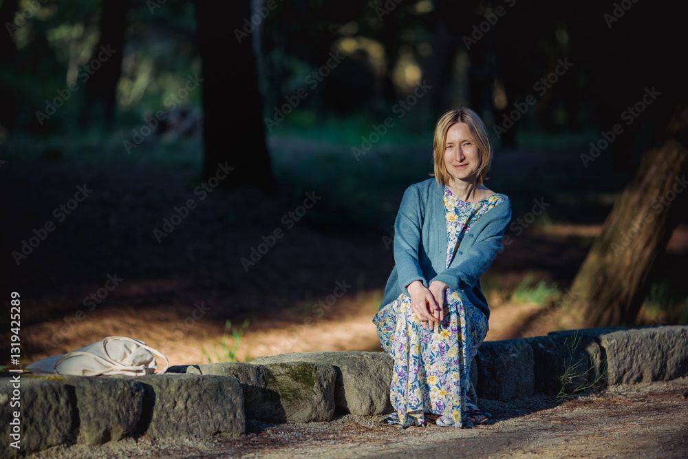 A woman sits on a stone-lined curb in a park during spring, surrounded by fresh greenery and blossoming nature.