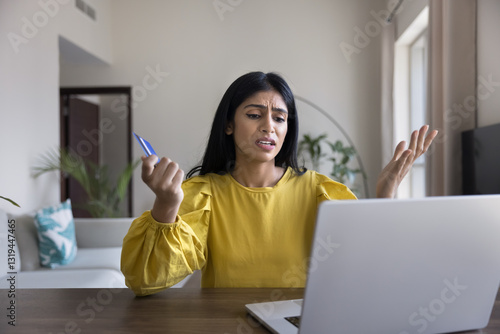 Indian woman looks at laptop screen with angry expression while holds card, try to make payment unsuccessfully, feels concerned due to additional fees or shipping costs during checkout, card limit
