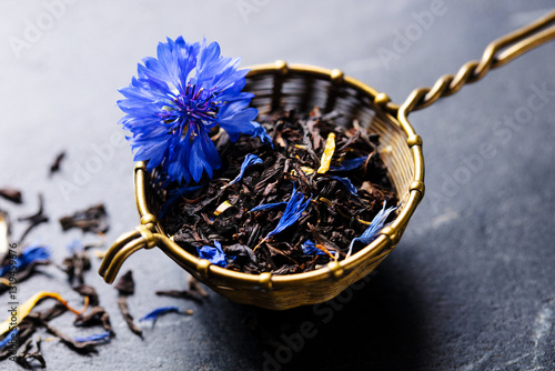Earl grey loose leaf tea blend with cornflower and orange blossom petals in golden in brass wire tea strainer. Marble background. Close up.