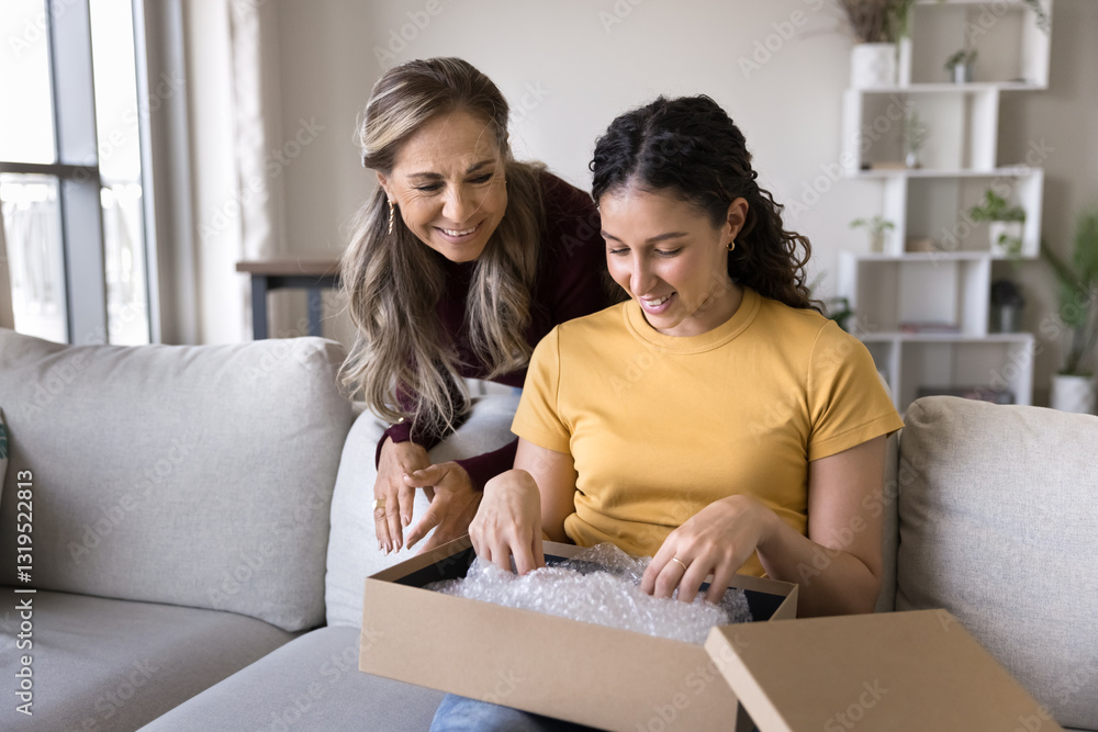 Happy Latina mother and her young adult daughter unpacking package together in living room, enjoy received parcel box contents with smile, feel satisfied with quality and express transporting services Happy Latina mother and her young adult daughter unpacking package together in living room, enjoy received parcel box contents with smile, feel satisfied with quality and express transporting services