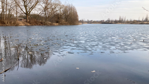 Early spring landscape featuring a partially frozen lake with a thin layer of melting ice. Bare trees line shore, reflecting on thawing surface. Transitioning from winter to spring.