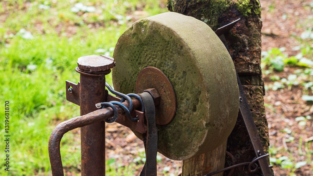 Rusty sharpening stone on a wooden pole in the woods. The sharpening stone is partially covered with moss and dirt. A metal rod with a handle, which was used for turning, comes off of it. Rusty sharpening stone on a wooden pole in the woods. The sharpening stone is partially covered with moss and dirt. A metal rod with a handle, which was used for turning, comes off of it.