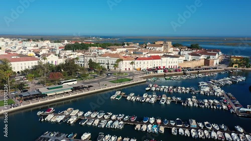 Aerial view of Faro, Portugal, featuring a marina with boats, a waterfront promenade, tree-lined streets, historic architecture, and the blue waters of the Ria Formosa lagoon