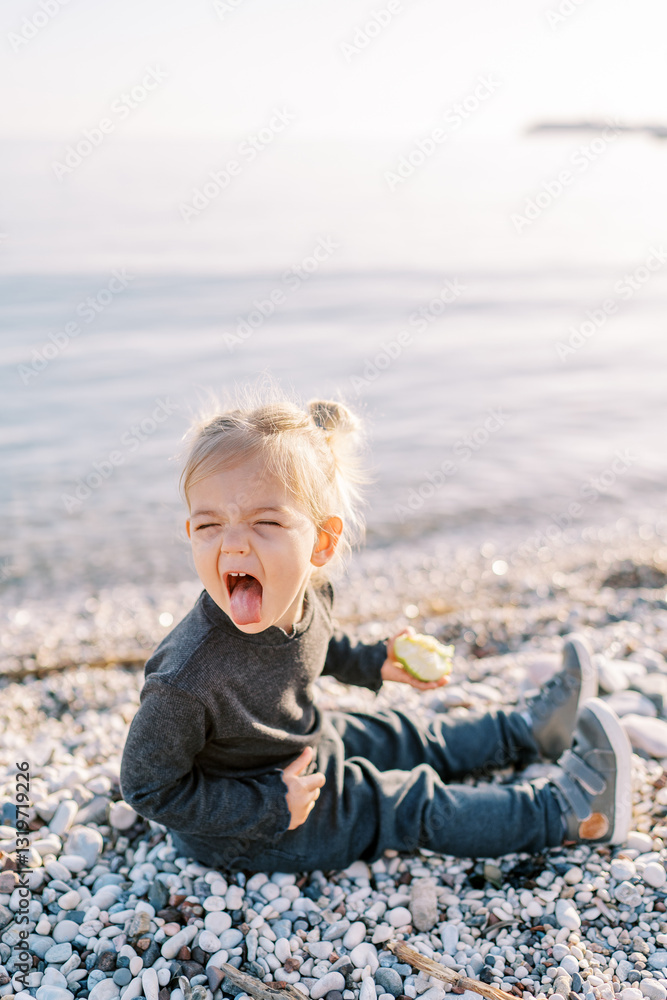 Little girl with an apple grimaces, sticking out her tongue while sitting on a pebble beach Little girl with an apple grimaces, sticking out her tongue while sitting on a pebble beach