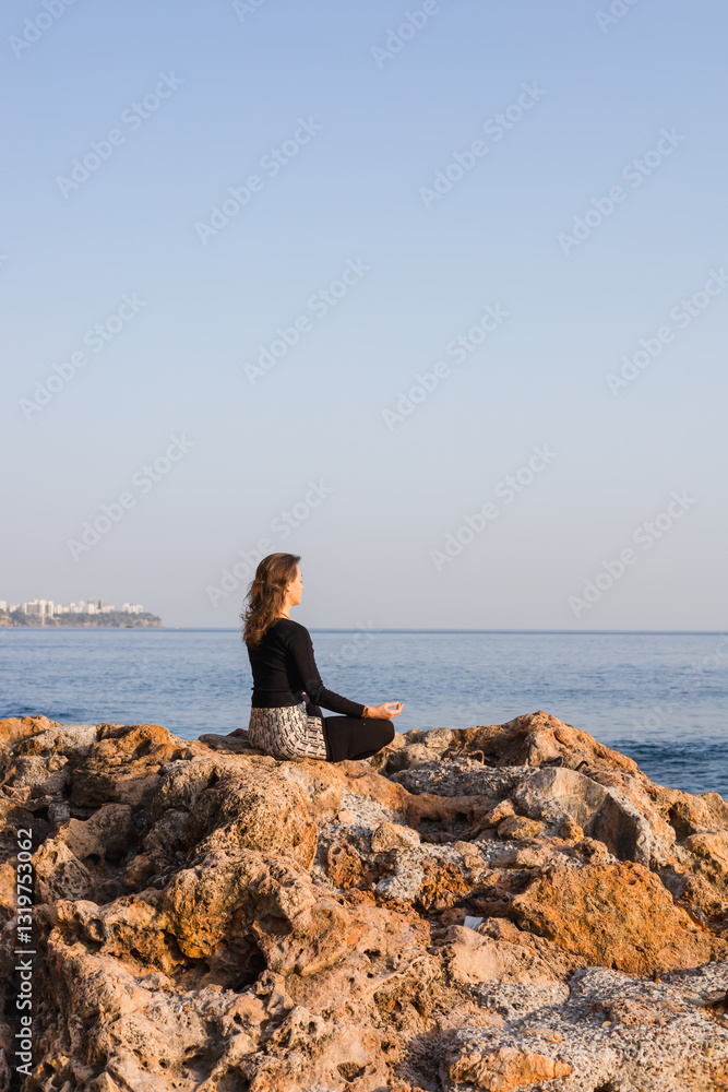 Young woman meditating on rocky beach with hands clasped sitting in lotus pose Young woman meditating on rocky beach with hands clasped sitting in lotus pose