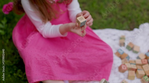 A boy and girl play with wooden alphabet blocks on a white blanket in the grass. The warm lighting and greenery evoke a nostalgic childhood moment, perfect for spring, summer, or Easter themes.