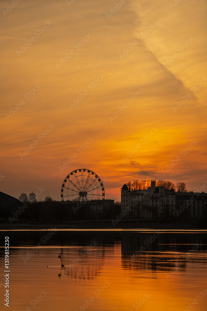 Obraz premium Latvian sunset with Riga sunset silhouette city scape. Calm small european city skyline of ferris wheel and modern building silhouettes of shadows in evening orange sun skies