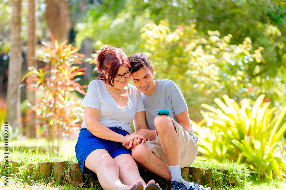 Fototapeta premium Happy adult son and mother sitting outdoors, holding hands and smiling while looking at a smartphone. Mother's Day and Mothering Sunday concept