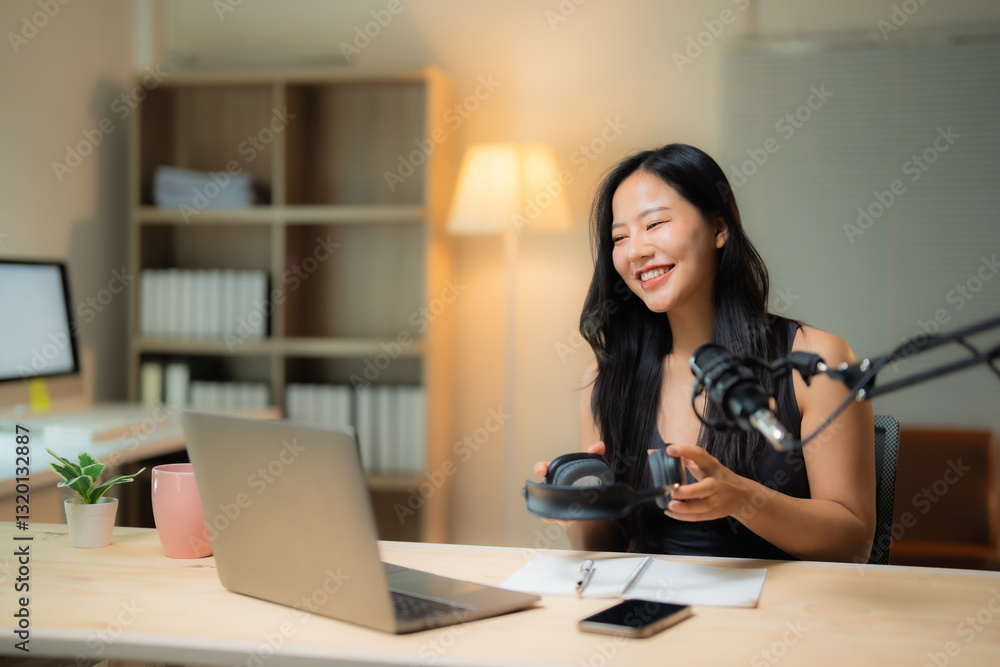 Happy podcaster preparing to record audio content in her home studio, holding headphones, smiling and looking at laptop, with microphone, notebook and smartphone on desk Happy podcaster preparing to record audio content in her home studio, holding headphones, smiling and looking at laptop, with microphone, notebook and smartphone on desk