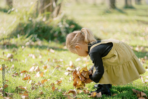 Schilderij op canvas Little girl collects a bouquet of fallen leaves, bending over on a sunny lawn