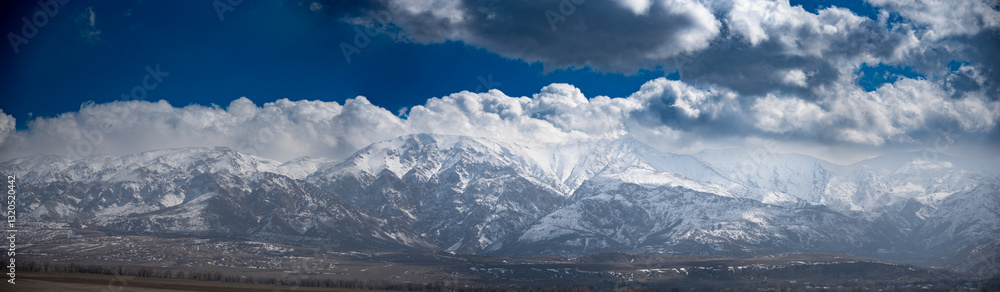 Obraz premium he image shows a majestic mountain range covered in snow under a dramatic sky filled with large, fluffy clouds. The mountains have a rugged and steep appearance, with various peaks and ridges visible.