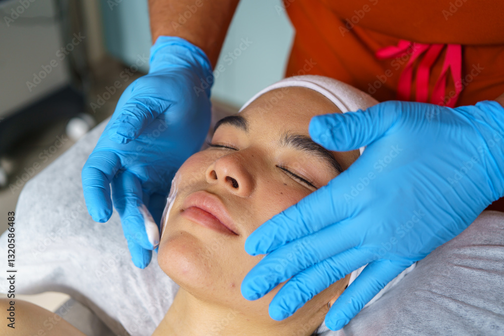 Beautician wearing blue gloves preparing face of young woman for ultrasonic facial cleansing by applying white cream Beautician wearing blue gloves preparing face of young woman for ultrasonic facial cleansing by applying white cream