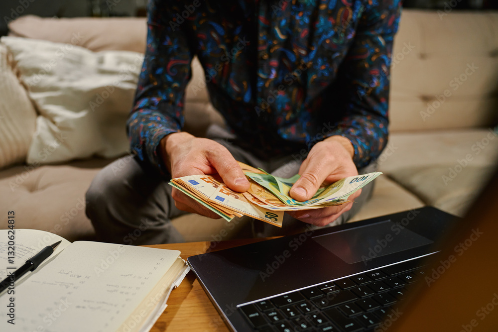 Man holding euro banknotes, sitting on sofa in front of table with laptop and notebook. Person counting remaining money after paying bills and utility expenses. Personal finance and home budget