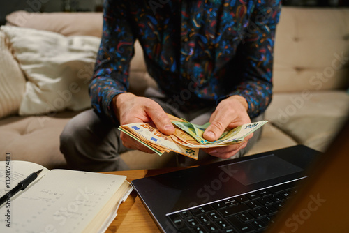 Obraz na płótnie Man holding euro banknotes, sitting on sofa in front of table with laptop and notebook. Person counting remaining money after paying bills and utility expenses. Personal finance and home budget w salonie