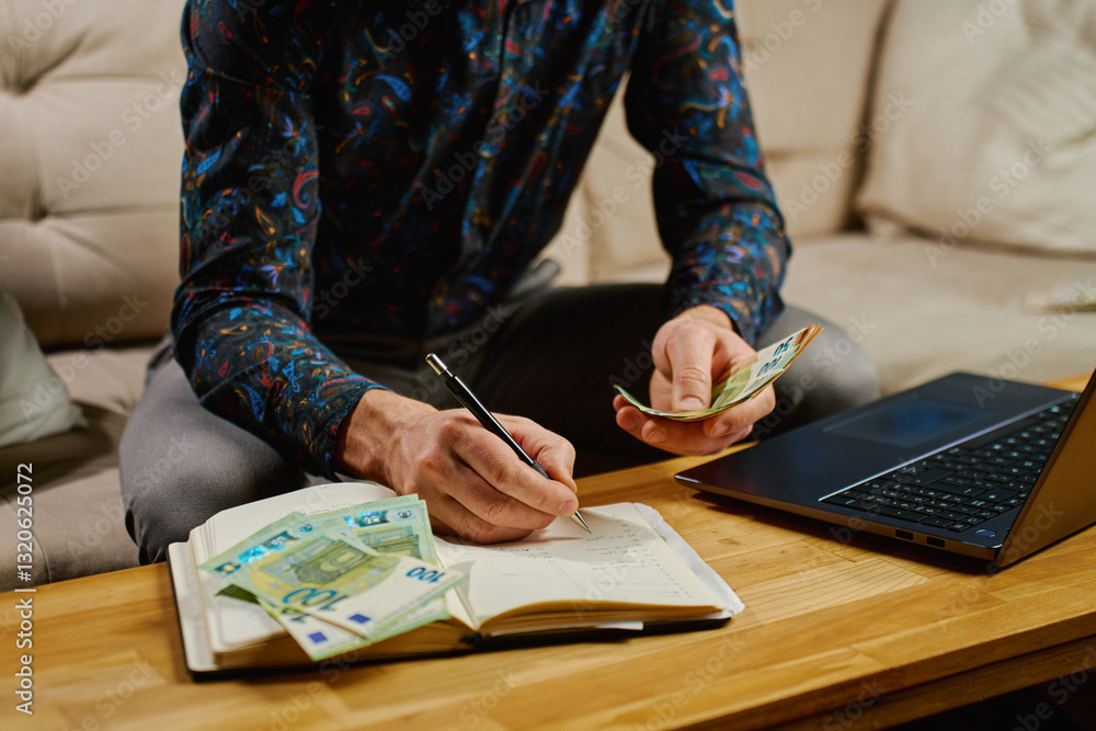 Man managing finances, writing in notebook while holding euro banknotes. Concept of budgeting, saving and financial planning